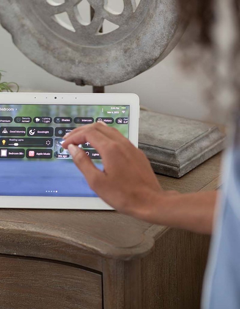A person using a tablet to control smart home devices, with a wooden piece of furniture and decorative object in the background.