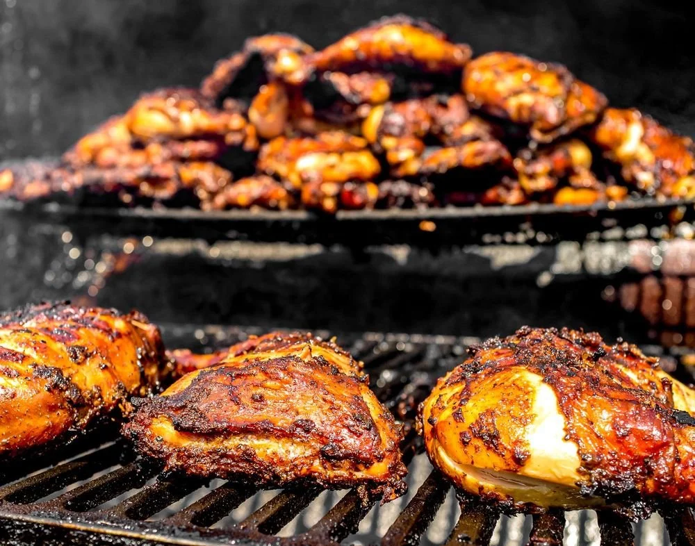 Close-up of spicy grilled chicken thighs and whole peppers on a barbecue grill, with a mound of grilled peppers in the background.