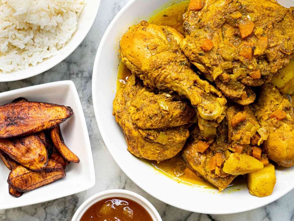 Plate of chicken cooked with vegetables in a yellow curry, a side of rice, grilled plantains, and sauce on a gray surface.