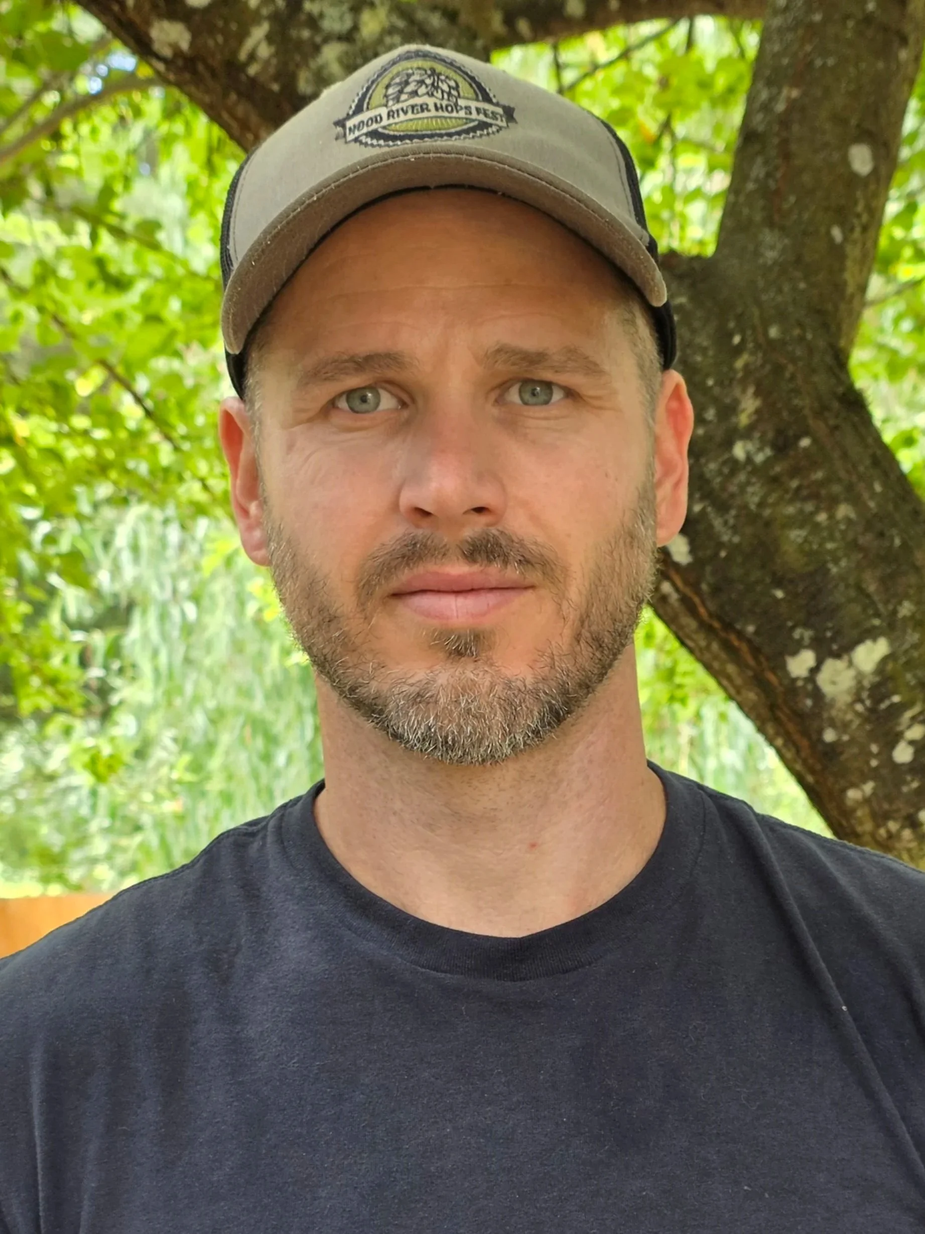 A close-up of a man with blue eyes, a beard, and short hair, wearing a black T-shirt and a baseball cap with a logo, standing outdoors with green leaves and a tree in the background.