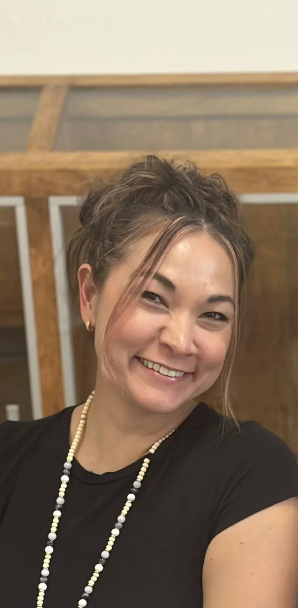 A smiling woman with short brown hair and a side-swept bang, wearing a black top and a beaded necklace, in a room with wooden and metal furniture.