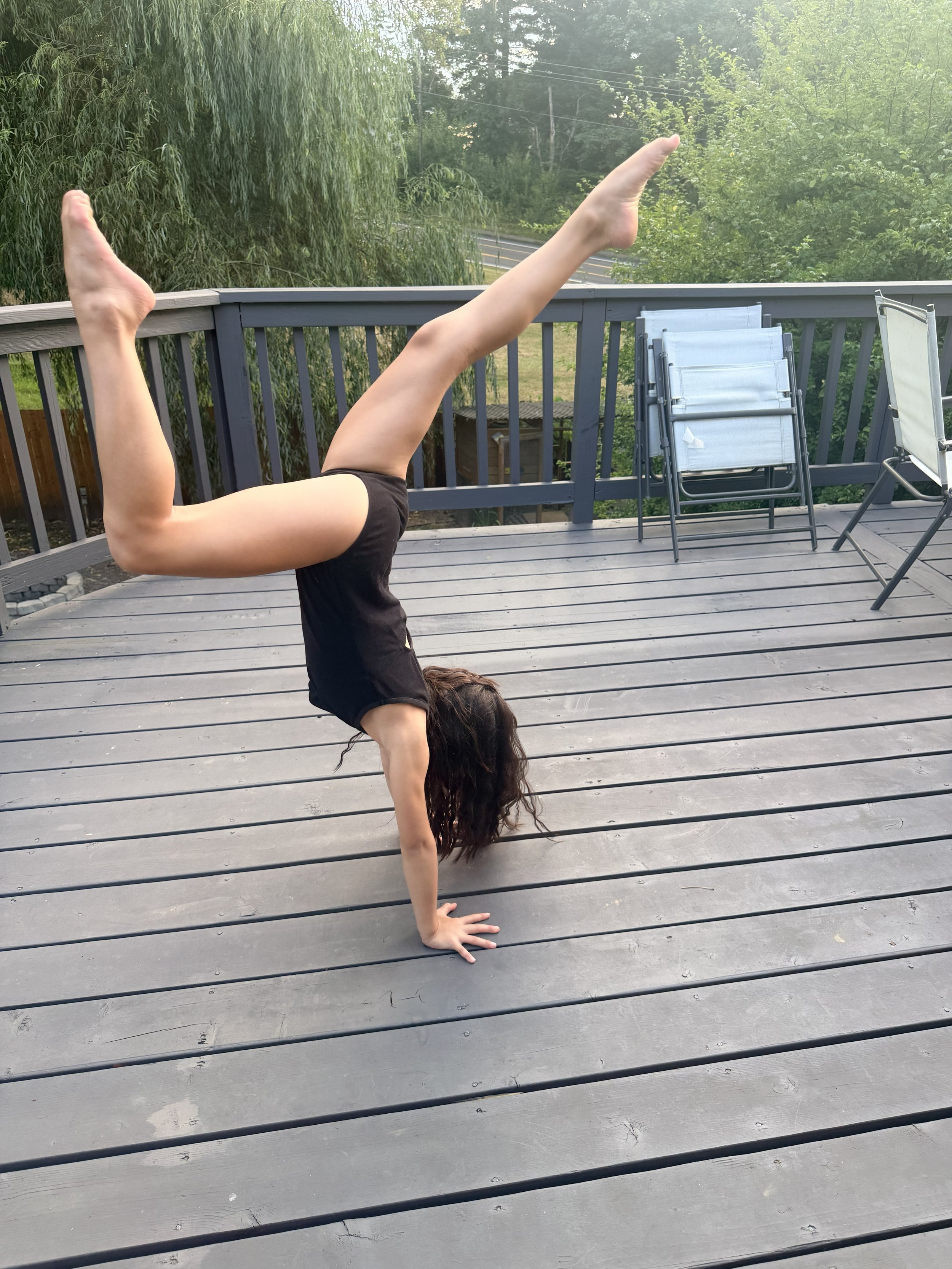 Person performing a handstand on a wooden deck outdoors, with their legs extended in a straight line. Folding chairs are stacked nearby, and lush greenery is visible in the background.