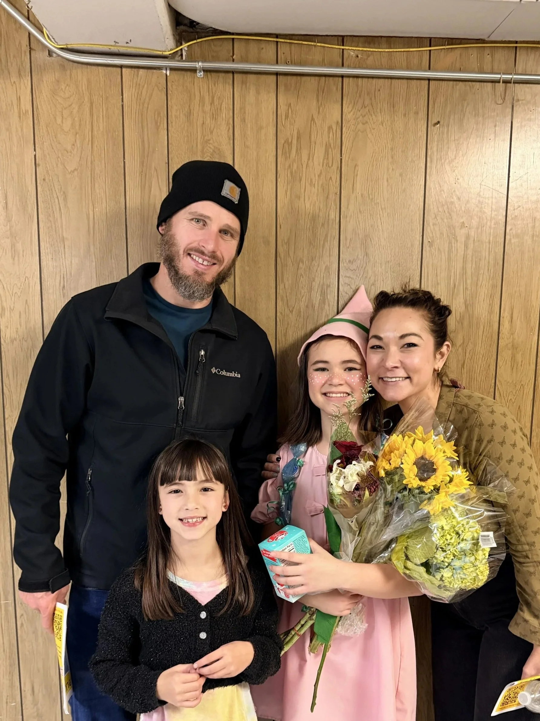 A woman in a tan jacket holding a bouquet of yellow flowers, a young girl in a pink dress with face paint, a man in a black jacket and black beanie, and a young girl with long dark hair holding a juice box, all smiling in front of a wood-paneled wall.