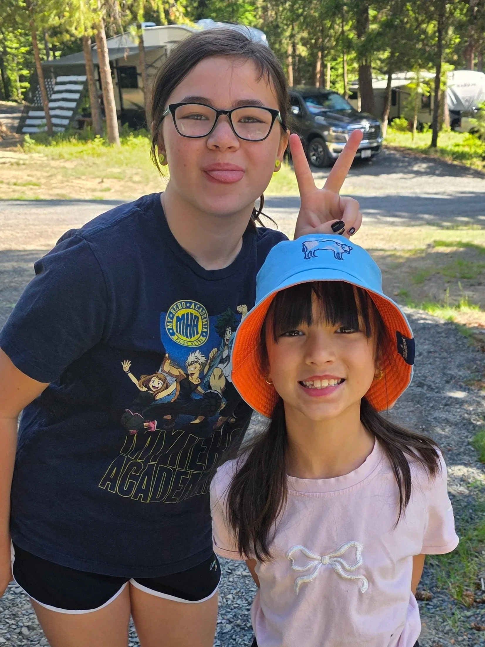 Two young girls outdoors in a wooded area, one making a peace sign with her hand. Both are smiling and dressed casually, with tents and vehicles in the background.