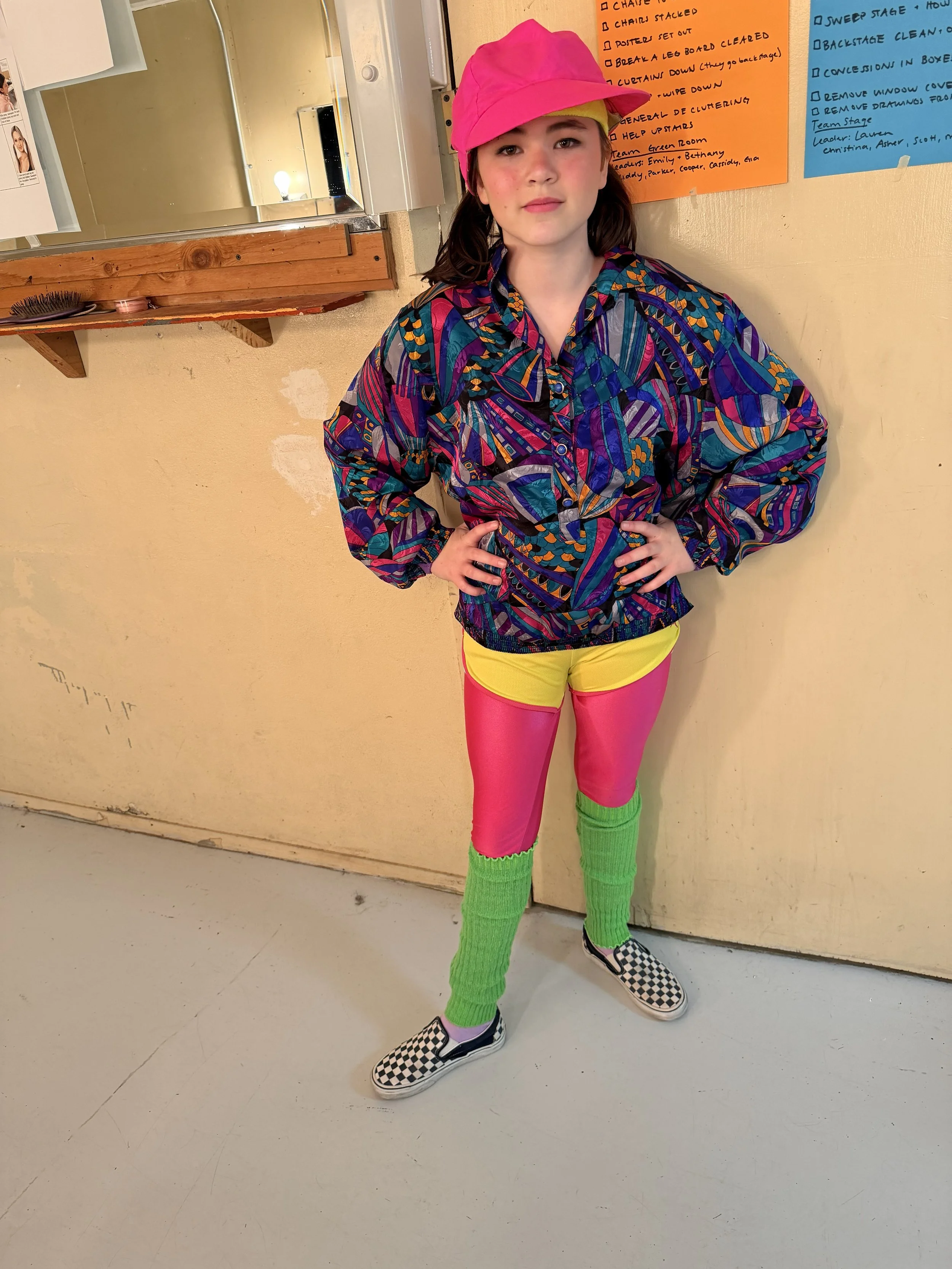 A young girl with brown hair, wearing a pink cap, colorful patterned jacket, bright pink leggings, green leg warmers, and black-and-white checkered slip-on shoes, standing against a beige wall with her hands on her hips.