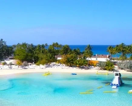 Tropical beach with clear blue water, palm trees, and boats near the shoreline, with the ocean in the background.
