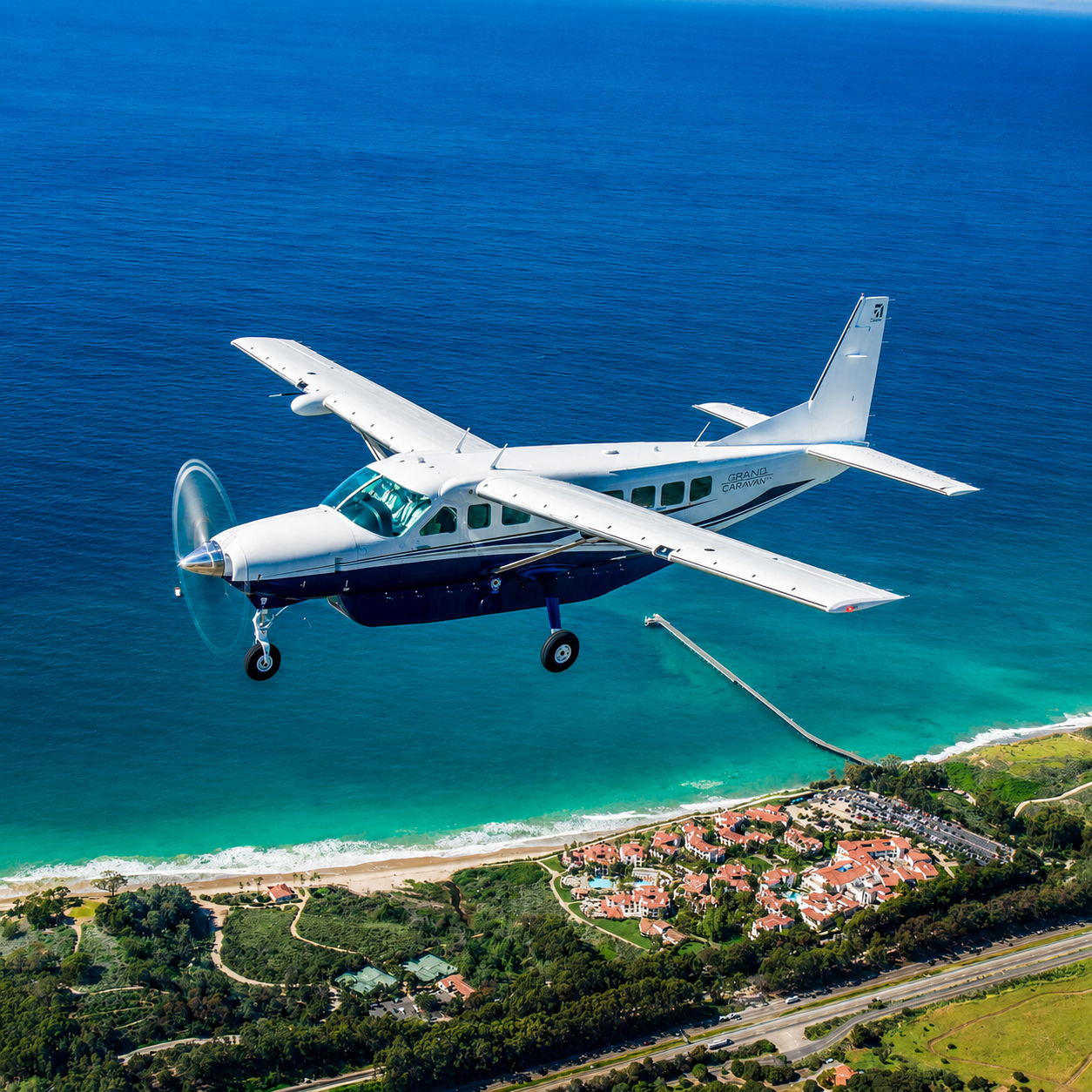 A small white and blue airplane flying over a beachside community with yellow sands, green vegetation, and a shoreline with turquoise water.