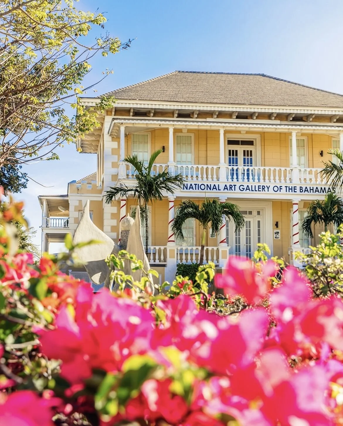The yellow building is the National Art Gallery of the Bahamas. It has a classic architectural style with a large balcony, white railing, and columns with red and white stripes. There are tropical palm trees, lush greenery, and pink flowers in the foreground, under a clear blue sky.