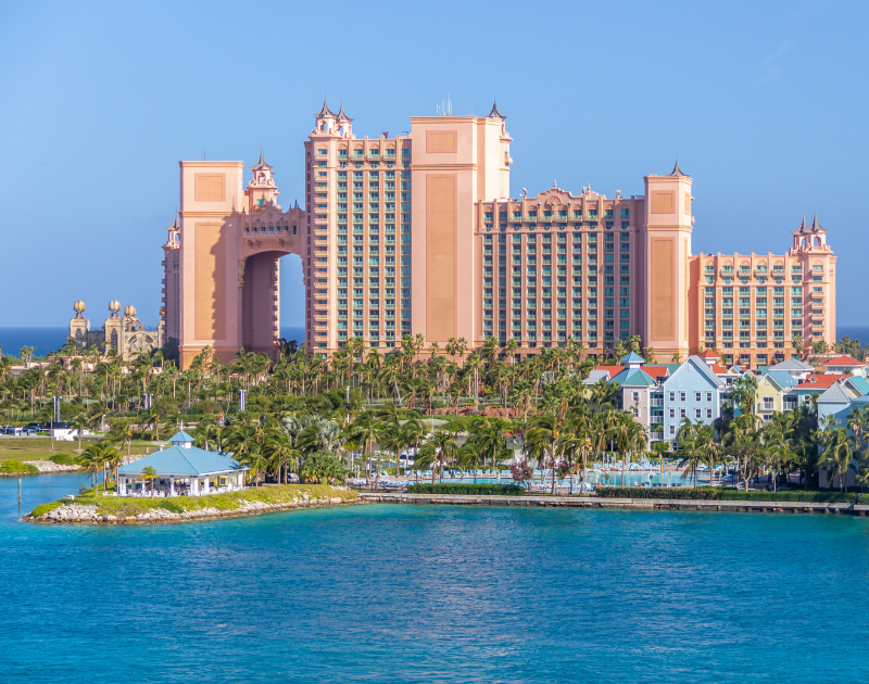 The image features a large pink resort hotel with a castle-like design, situated by a body of water with a marina, palm trees, and smaller colorful houses in the foreground.