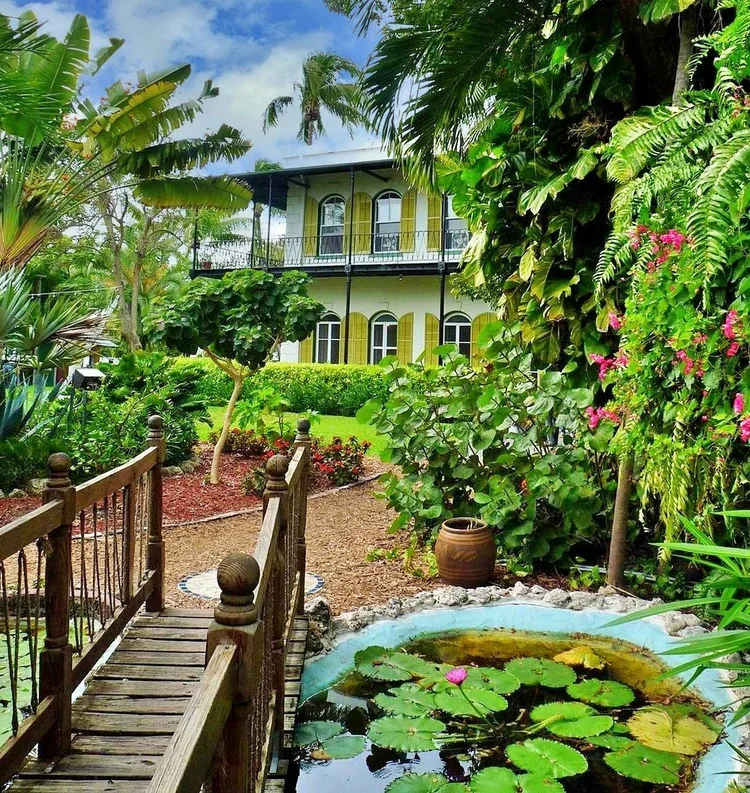A lush tropical garden with various plants, flowers, and trees. A small pond with lily pads and a pink water lily. A wooden footbridge crosses over the pond leading to a pathway. In the background, there is a yellow two-story house with arched windows and a balcony, surrounded by well-maintained green landscaping.