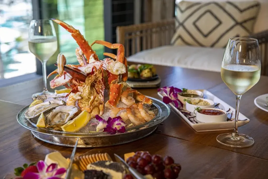 Seafood platter featuring lobster, crab, shrimp, and oysters with lemon wedges, served on ice. Two glasses of white wine and an assortment of dipping sauces and condiments on the side, with a cozy indoor setting and decorative pillows in the background.