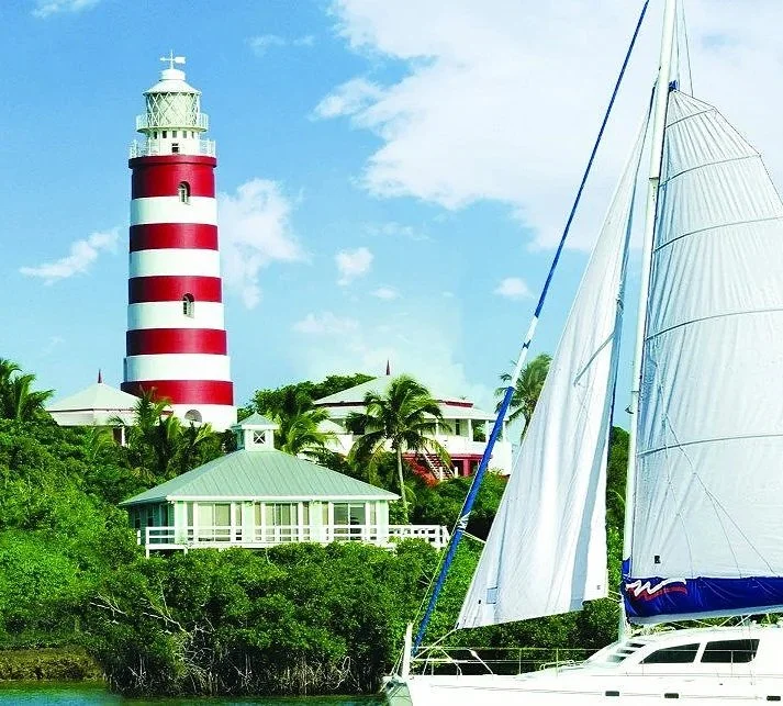 A sailboat with white sails in front of a lighthouse with red and white stripes, surrounded by palm trees and coastal buildings.