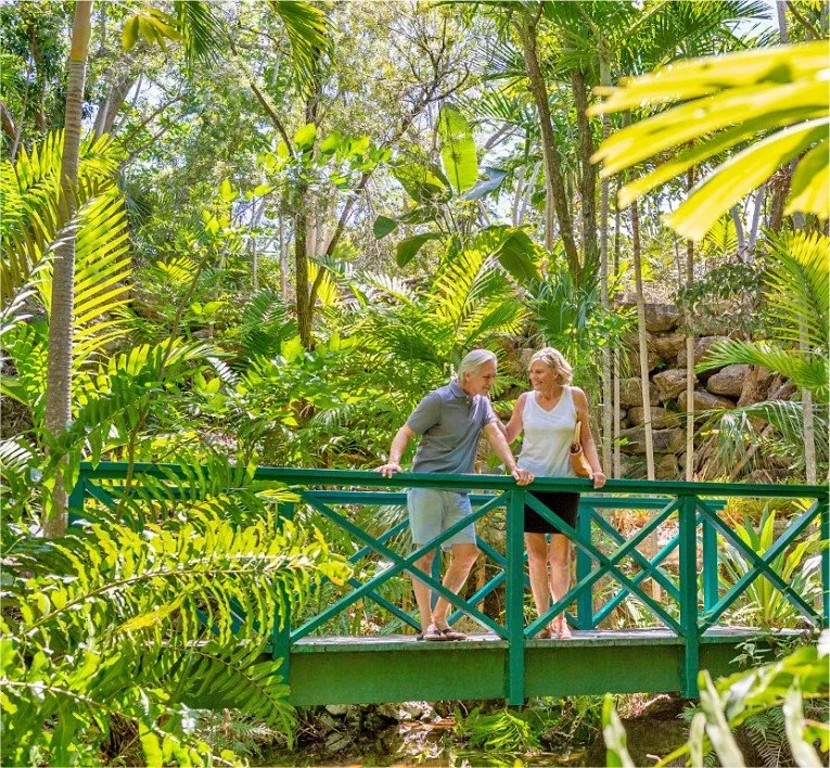 Two older women strolling on a small green bridge in a lush tropical garden, holding hands and smiling.