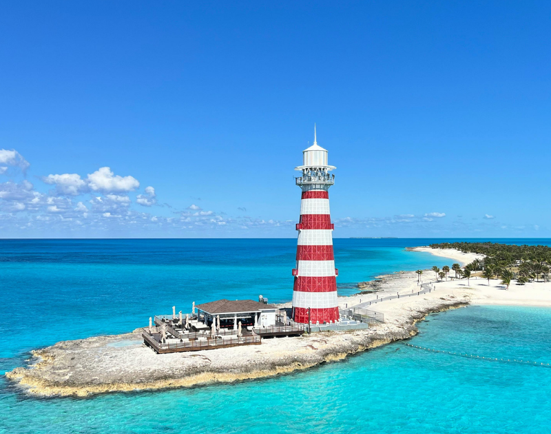 A red and white striped lighthouse on a small rocky island in the ocean, with a beach and trees in the background under a clear blue sky.