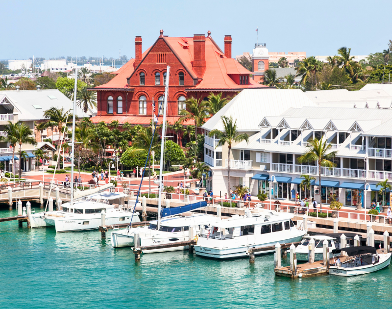 Marina with several boats docked, colorful buildings, palm trees, and a large historic red brick building in a waterfront town.