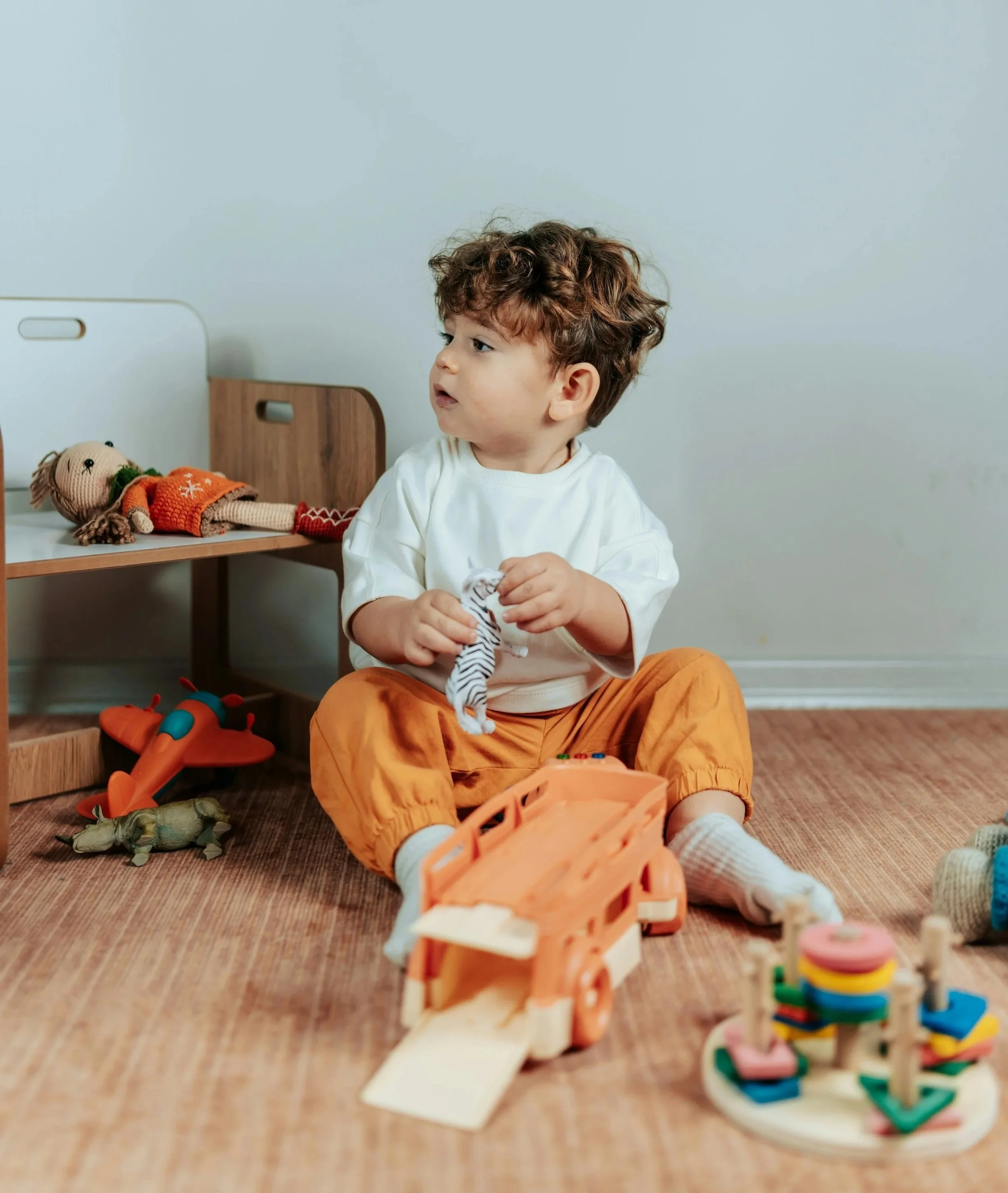 A young boy with curly brown hair sitting on the wooden floor, holding a small plush toy, working on expressive and receptive language skills during speech therapy for children in Bethesda, MD