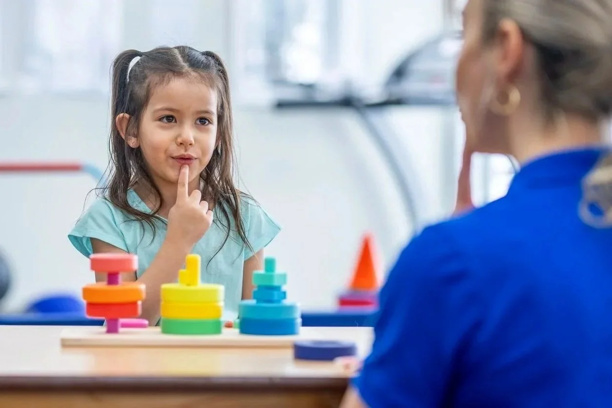 A young girl sitting across from a speech therapist, playing with brightly colored rings. The girl has her finger on her chin, working on decreasing a tongue thrust during myofunctional therapy for kids in Bethesda, MD.