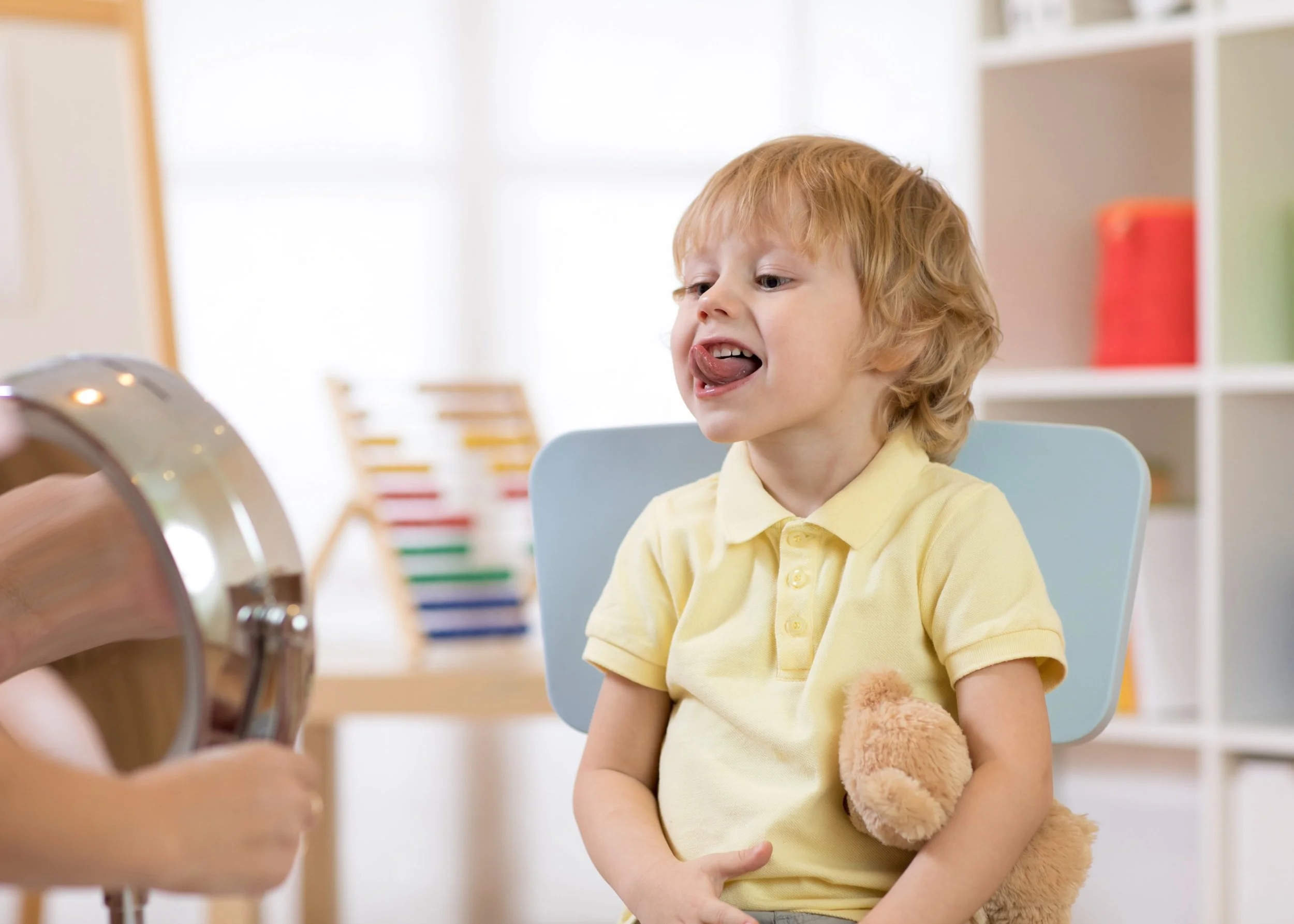 Young boy with red hair and freckles sitting at a table in a brightly lit room, sticking out his tongue, working on exercises for tongue tie during myofunctional therapy in Bethesda, MD