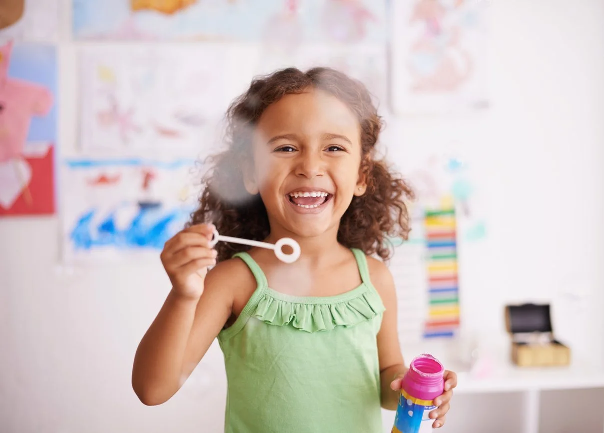 A young girl with curly hair wearing a green top, smiling and blowing bubbles in a brightly decorated room, working on speech and oral motor skills during speech therapy for children in Bethesda, MD