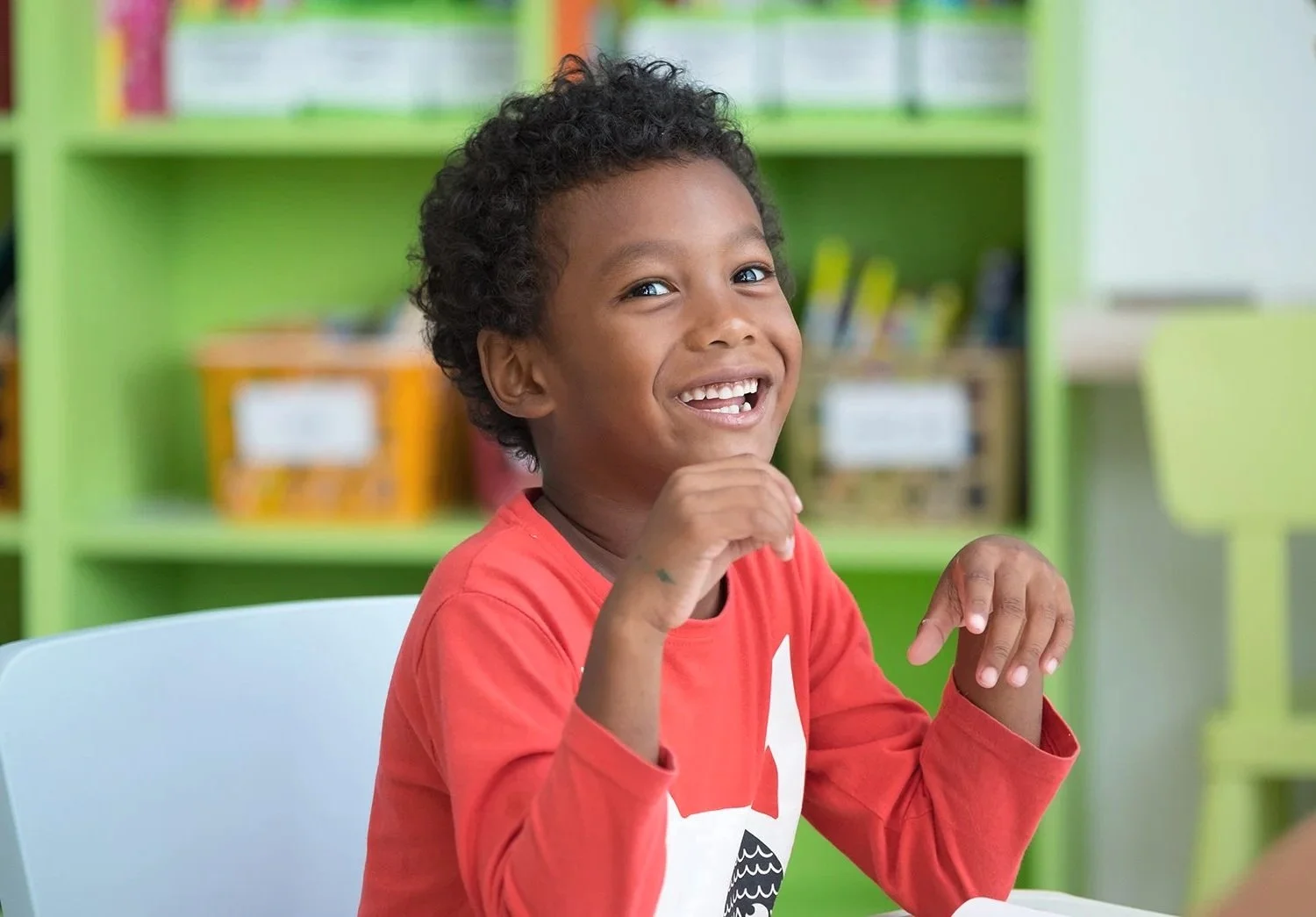 A young boy with curly hair wearing a red shirt, smiling happily, working on improving his speech sounds during speech therapy for a lisp in Bethesda, MD