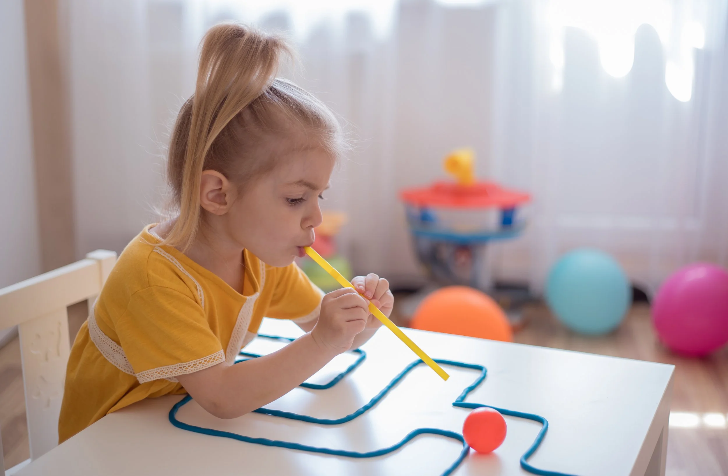 A young girl with blonde hair and a yellow shirt is sitting at a white table, using a straw to control the movement of a ball during myofunctional therapy for children in Bethesda, MD