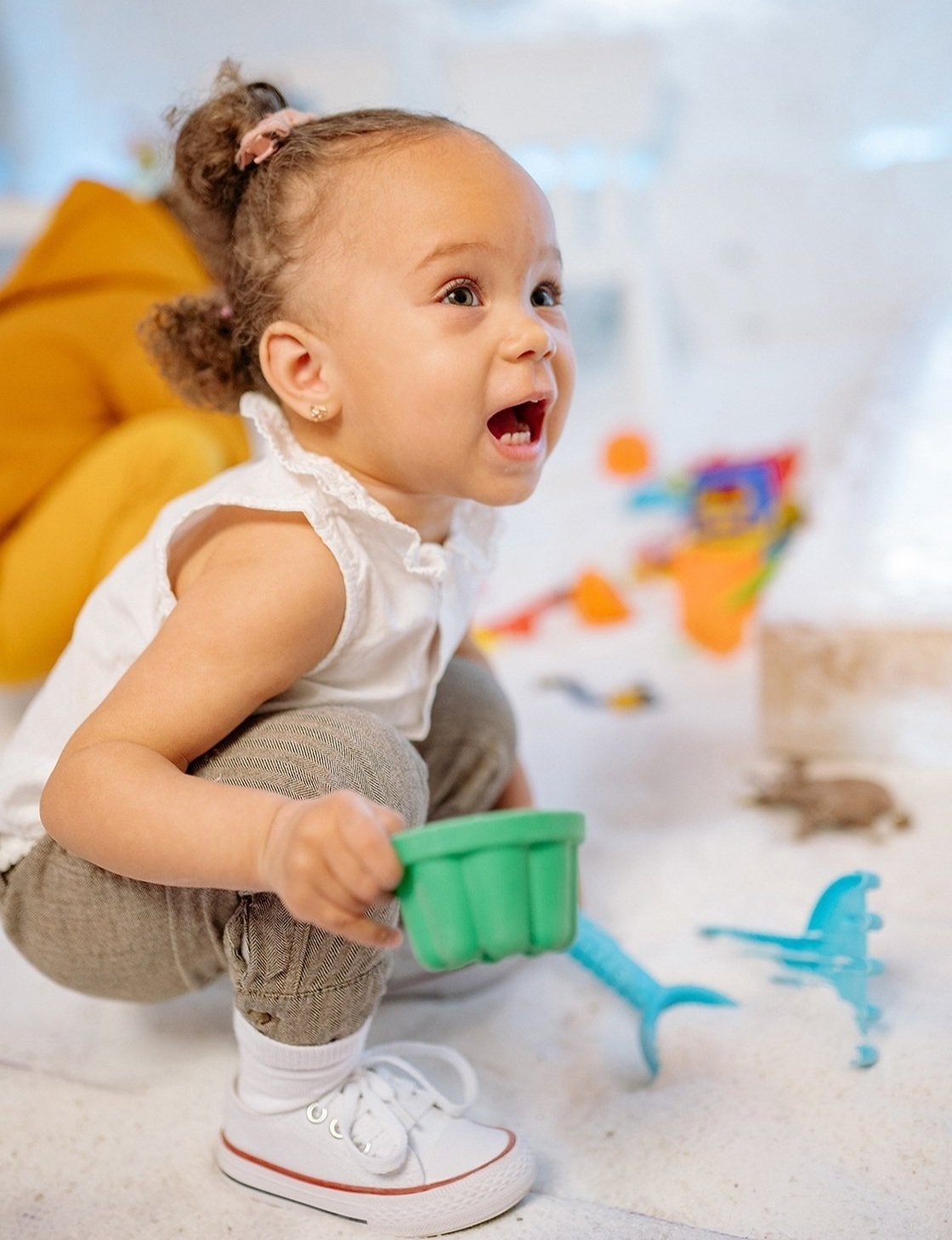 A young girl with curly hair tied in a bun, wearing a white sleeveless top and beige pants, squatting on the floor and playing with a green toy bucket and sand toys during language and communication therapy for late talkers in Bethesda, MD