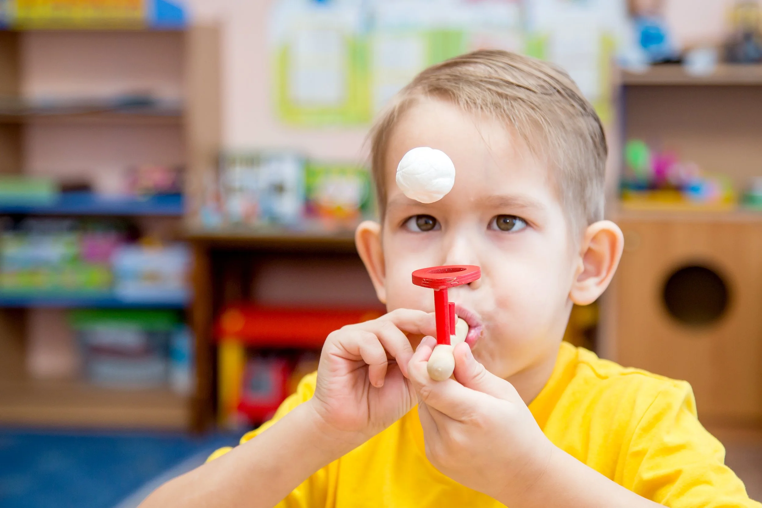 A young boy wearing a yellow shirt, using a blowing tool to hold a small ball up in the air while working on improved breathing and oral motor skills during myofunctional therapy for kids in Bethesda, MD