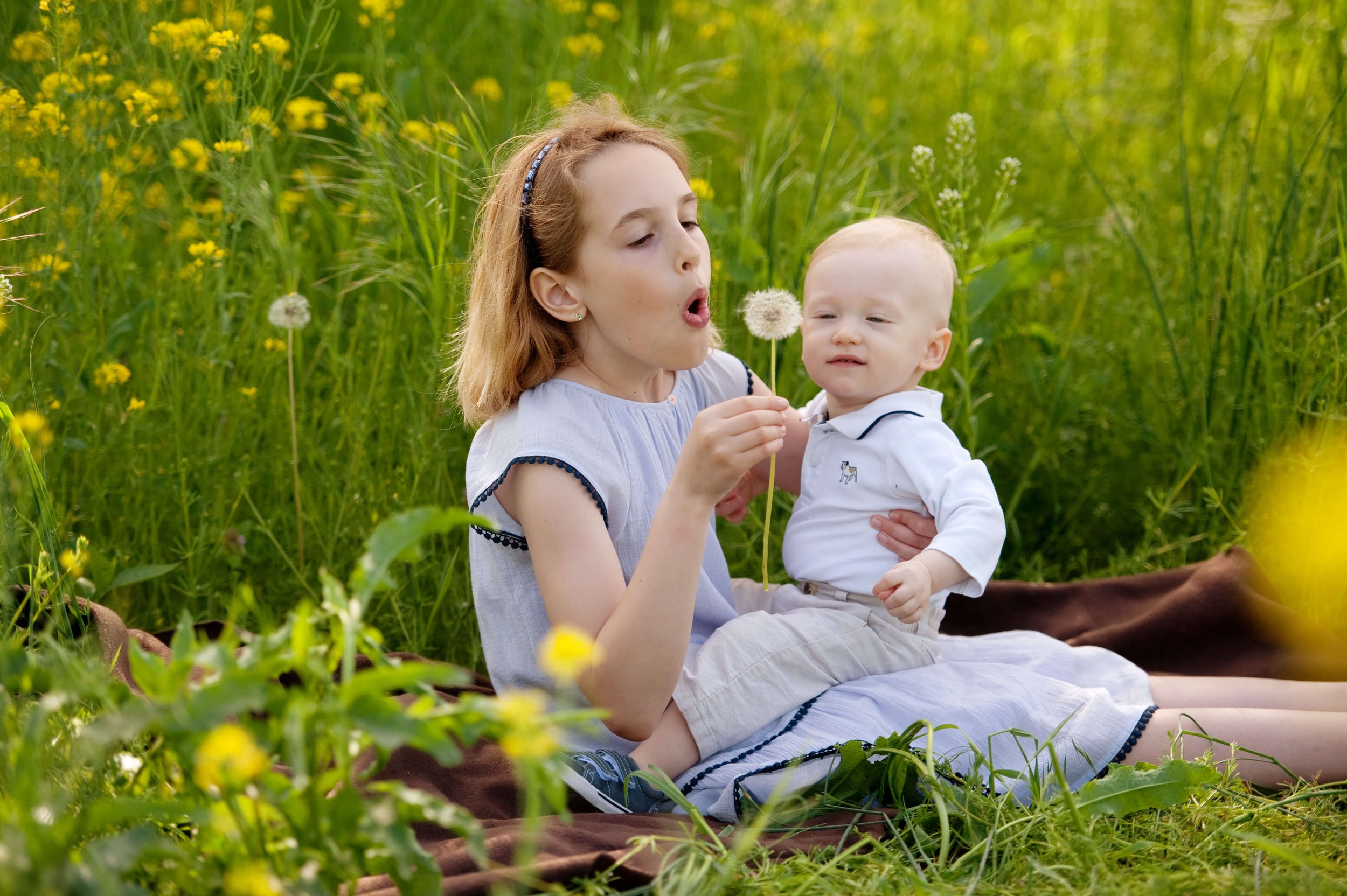 A young girl with light brown hair and a headband, sitting in a grassy field with yellow flowers, holds a dandelion and blows on it, while a baby with blond hair and a white shirt sits on her lap. Background for Thrive Speech and Wellness