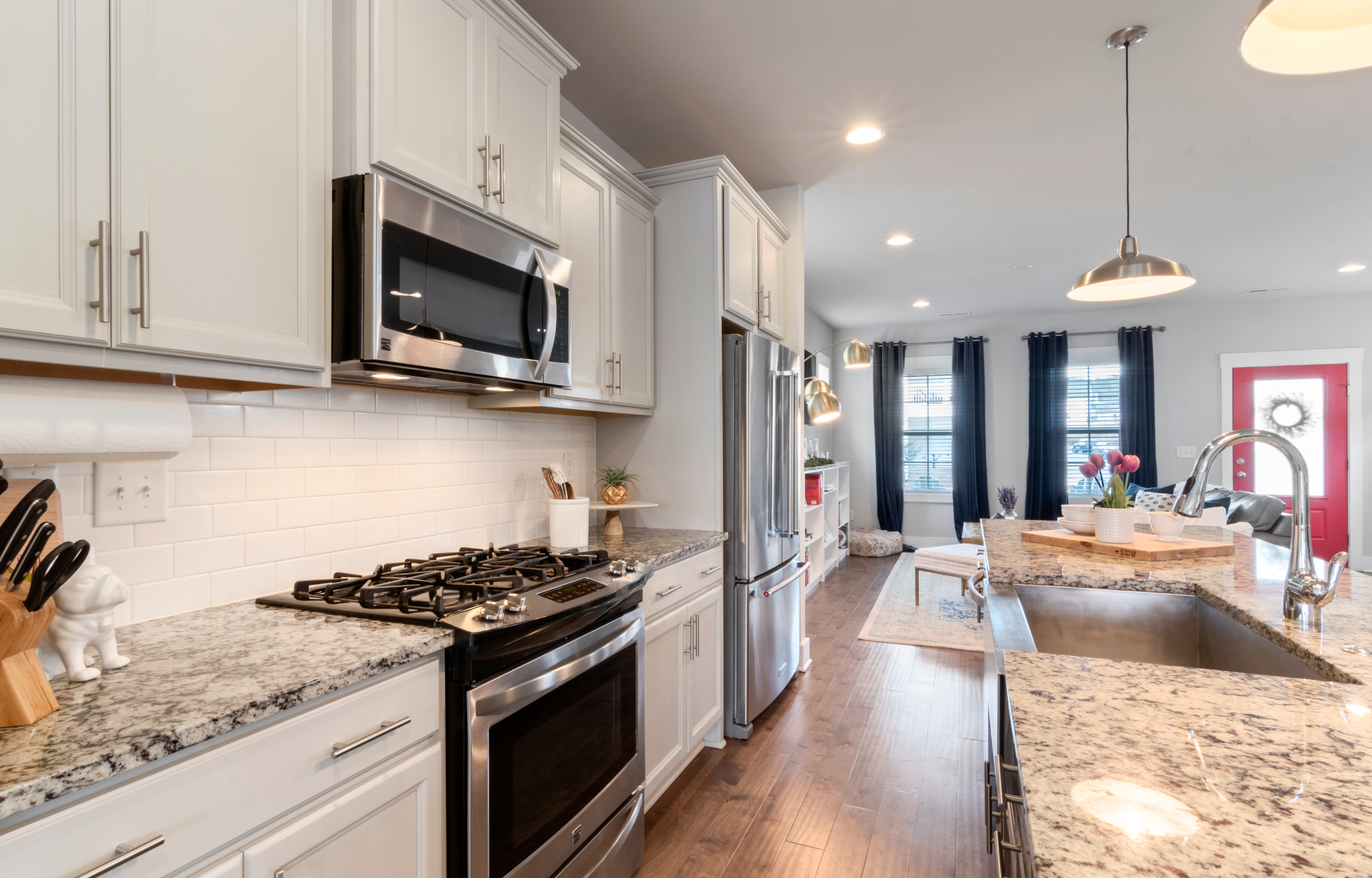 Modern kitchen with white cabinets, stainless steel appliances including a microwave and refrigerator, granite countertops, a gas stove, and a breakfast bar with a potted plant and bowls. The open living space has large windows with dark curtains, a sitting area with a red door, and hardwood flooring.