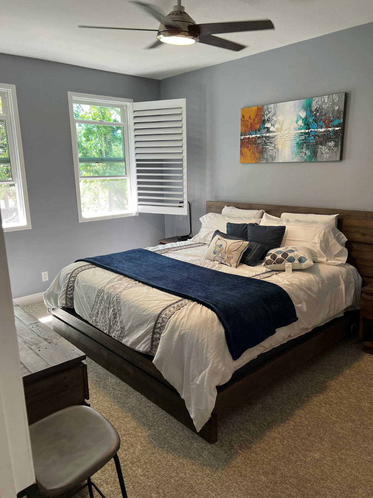 A bedroom with a bed covered in white and patterned bedding, decorated with multiple pillows, and a dark blue throw blanket. There are two windows with white shutters, a wooden nightstand, and a modern ceiling fan. An abstract colorful painting hangs above the bed, and a brown chair is partially visible in the foreground.