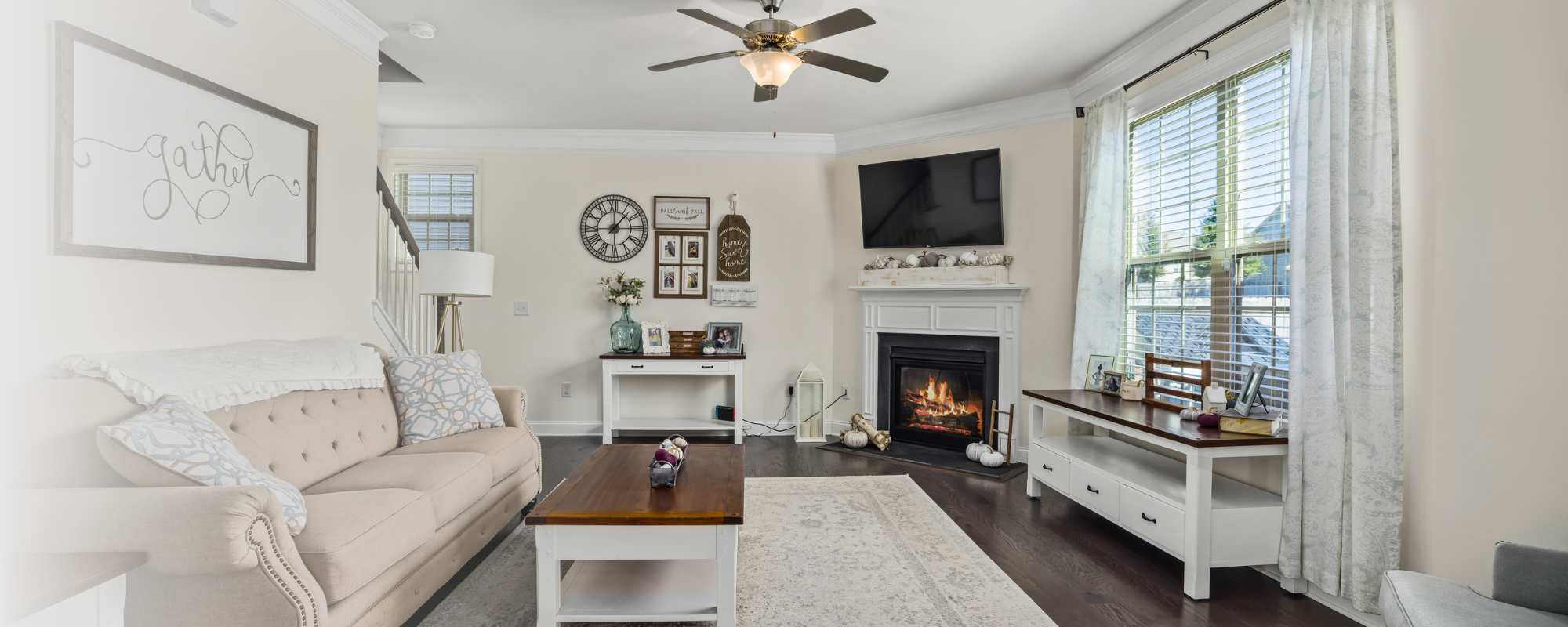 Living room with a beige couch, a coffee table, a white fireplace, a wall-mounted TV, and windows with white curtains, decorated for autumn with pumpkins and fall-themed decor.
