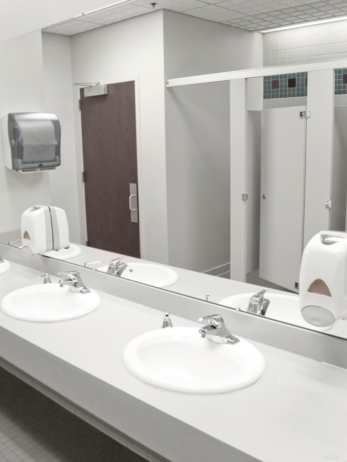 Public restroom with three sinks, soap dispensers, and mirror, with bathroom stalls in the background.