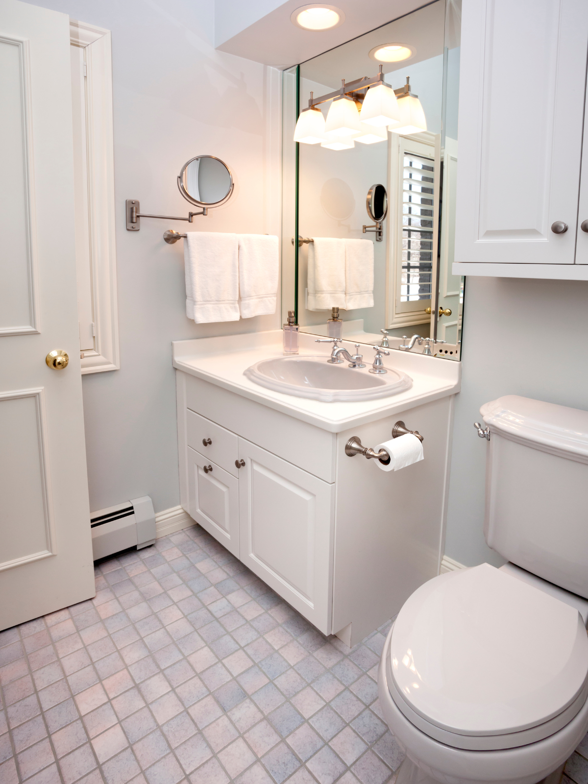 A small bathroom with a white vanity, a sink, a large mirror, a wall-mounted light fixture, white towels, and a toilet. The floor is tiled in light-colored square tiles. There is a window with shutters visible in the mirror.