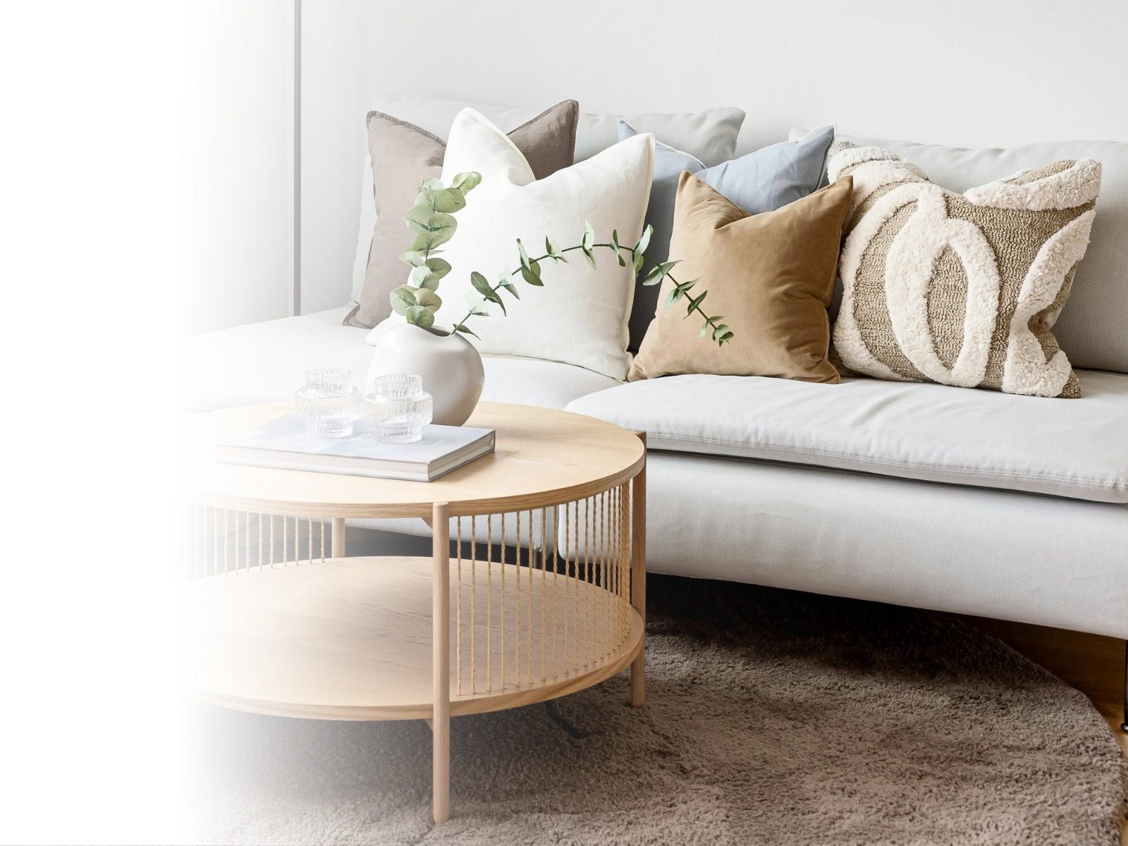 Living room with a white sofa, multiple neutral-colored pillows, a round wooden coffee table with a white vase and greenery, and a beige rug.