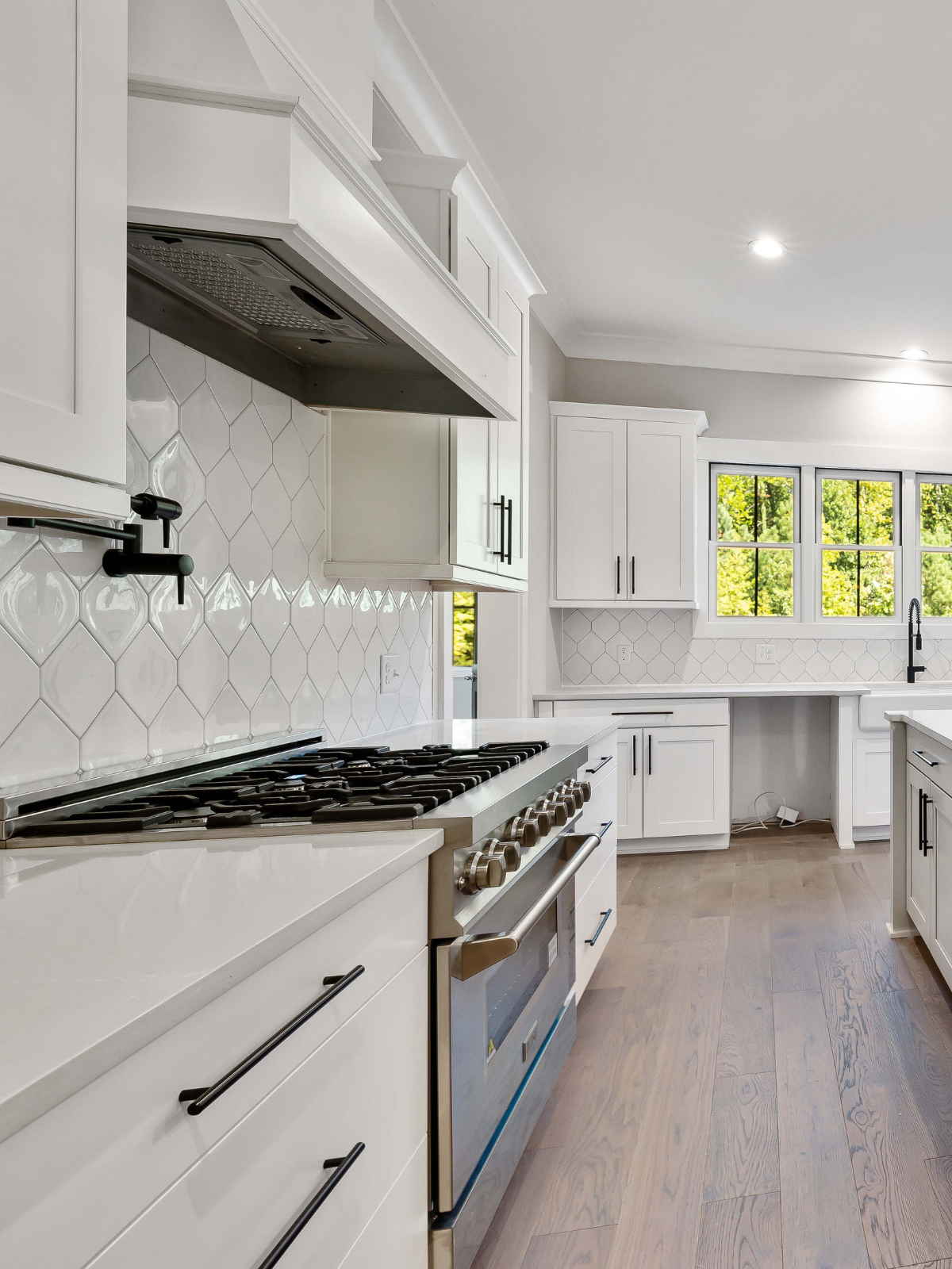 Modern white kitchen with wood flooring, gas stove, white cabinets, subway tile backsplash, and a window showing green trees outside.
