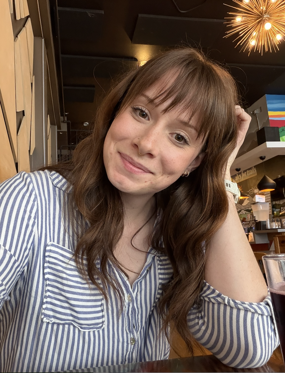 A young woman with long, wavy brown hair and a nose piercing, smiling in a restaurant or cafe. She is wearing a blue and white striped shirt and has a glass of dark beverage in front of her.