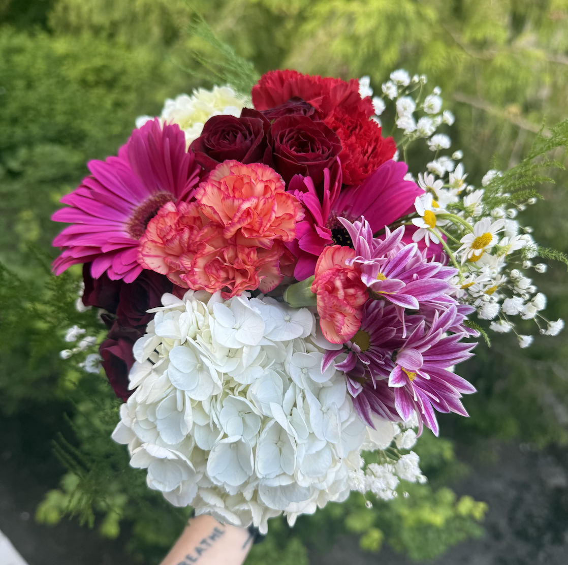 A colorful bouquet of flowers with pink, red, white, and purple blooms, held against a green outdoor background.