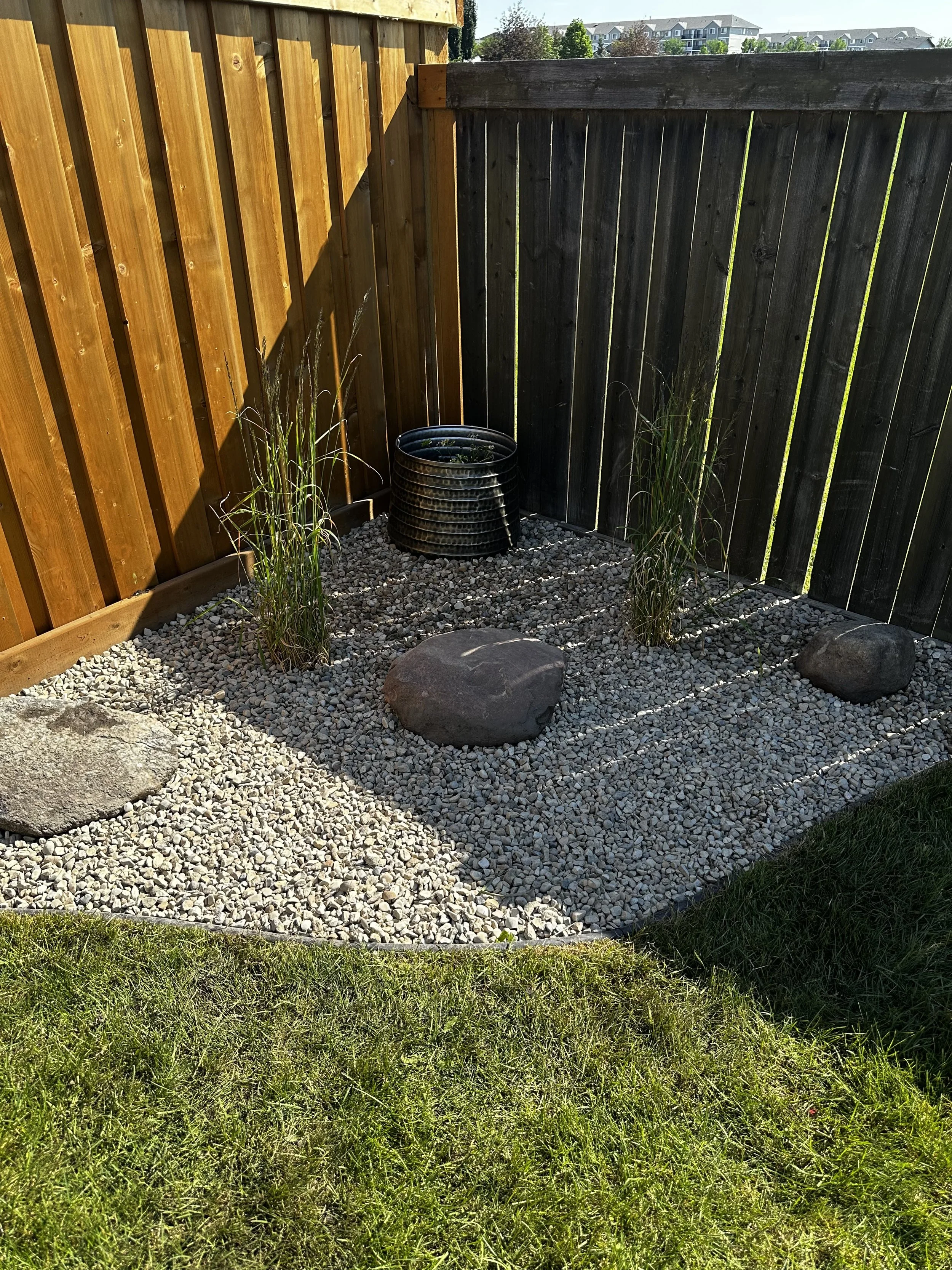 A small outdoor garden area with gravel ground cover, two tall ornamental grasses, a large flat stone, and a black metal decorative container in front of a wooden fence with and a dark colored fence adjacent, with a background of residential buildings.