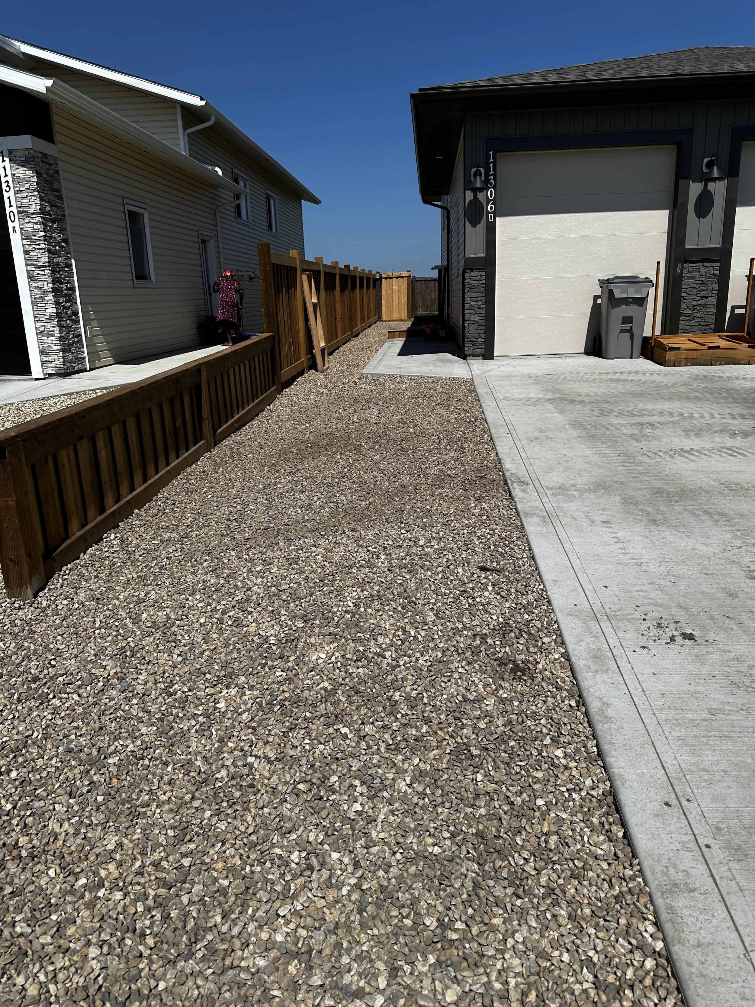 A narrow alleyway between two modern houses, showing a gravel pathway on the left, a paved driveway on the right, and a wooden fence at the back. A woman is walking near the house on the left, and a trash bin is placed in front of the garage on the right.