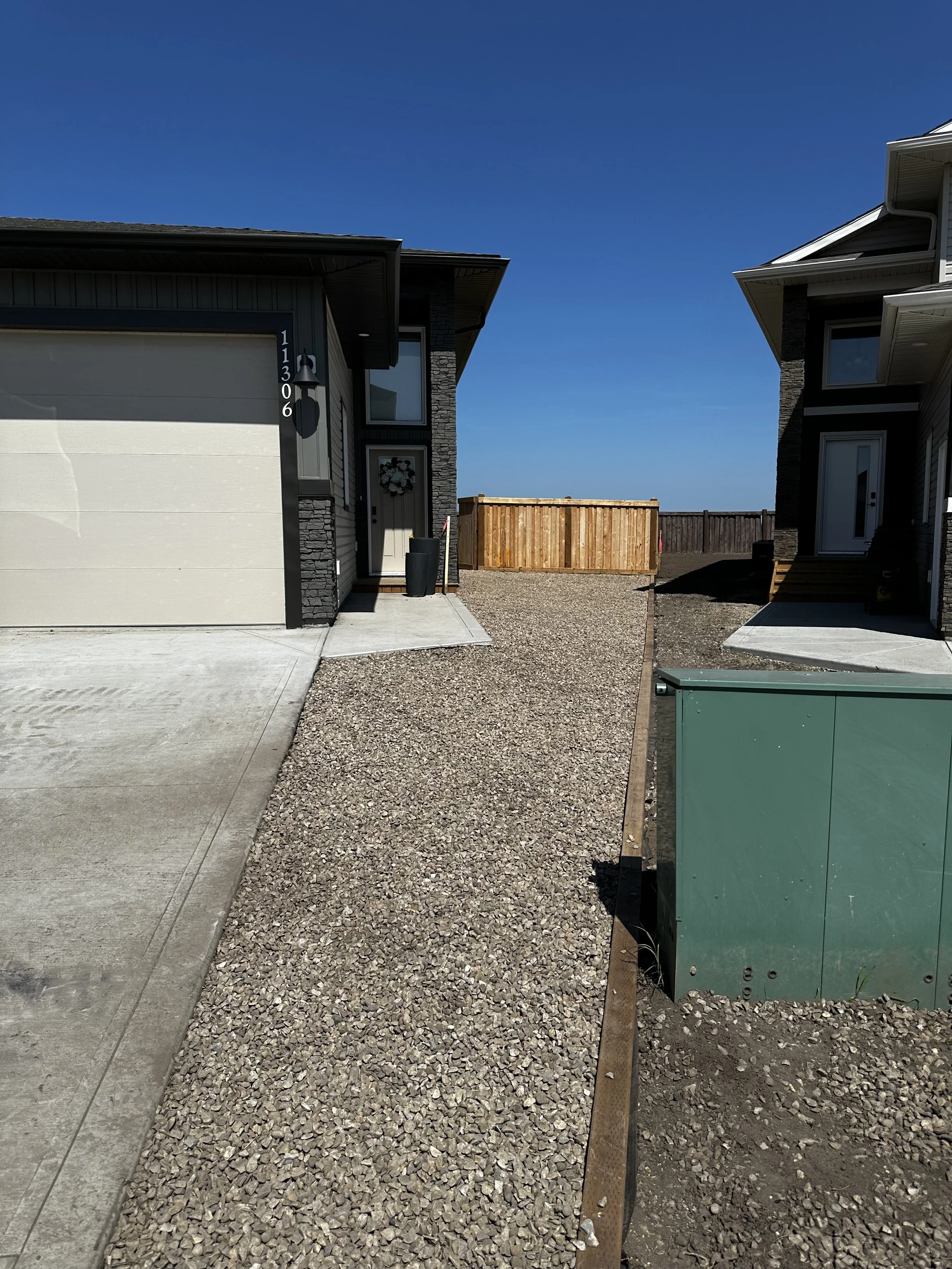 Newly constructed residential houses with a driveway, gravel yard, wooden fence, and utility box under a clear blue sky.