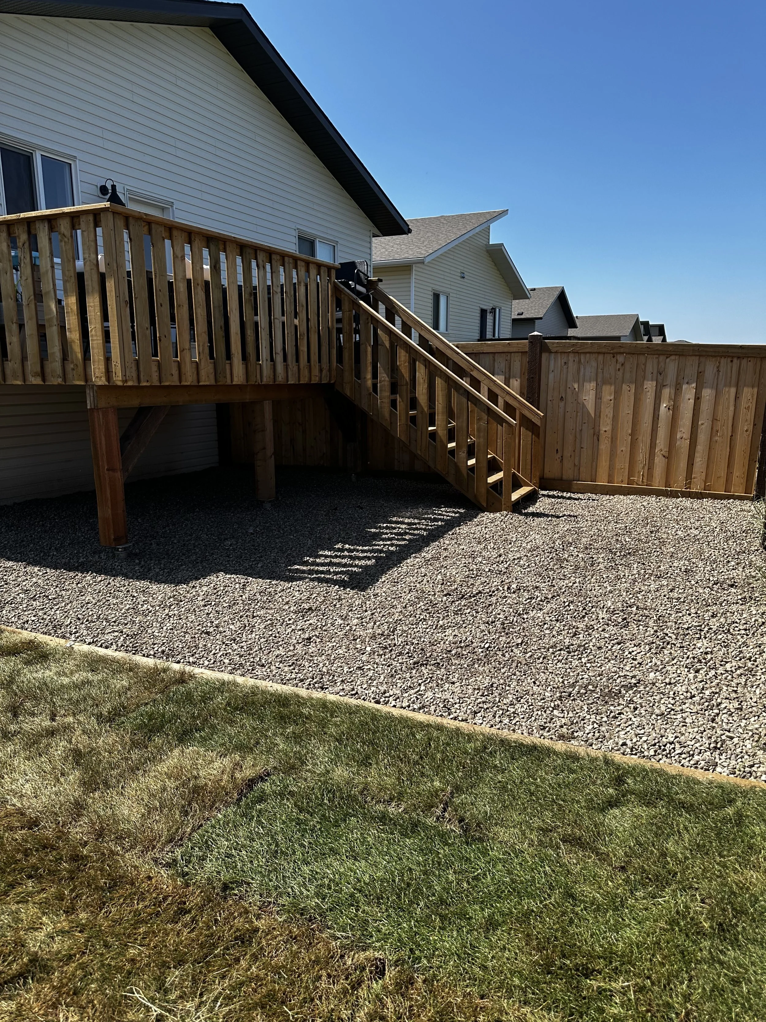 Backyard with a wooden deck, stairs, and a privacy fence, with neighboring houses and a clear blue sky.