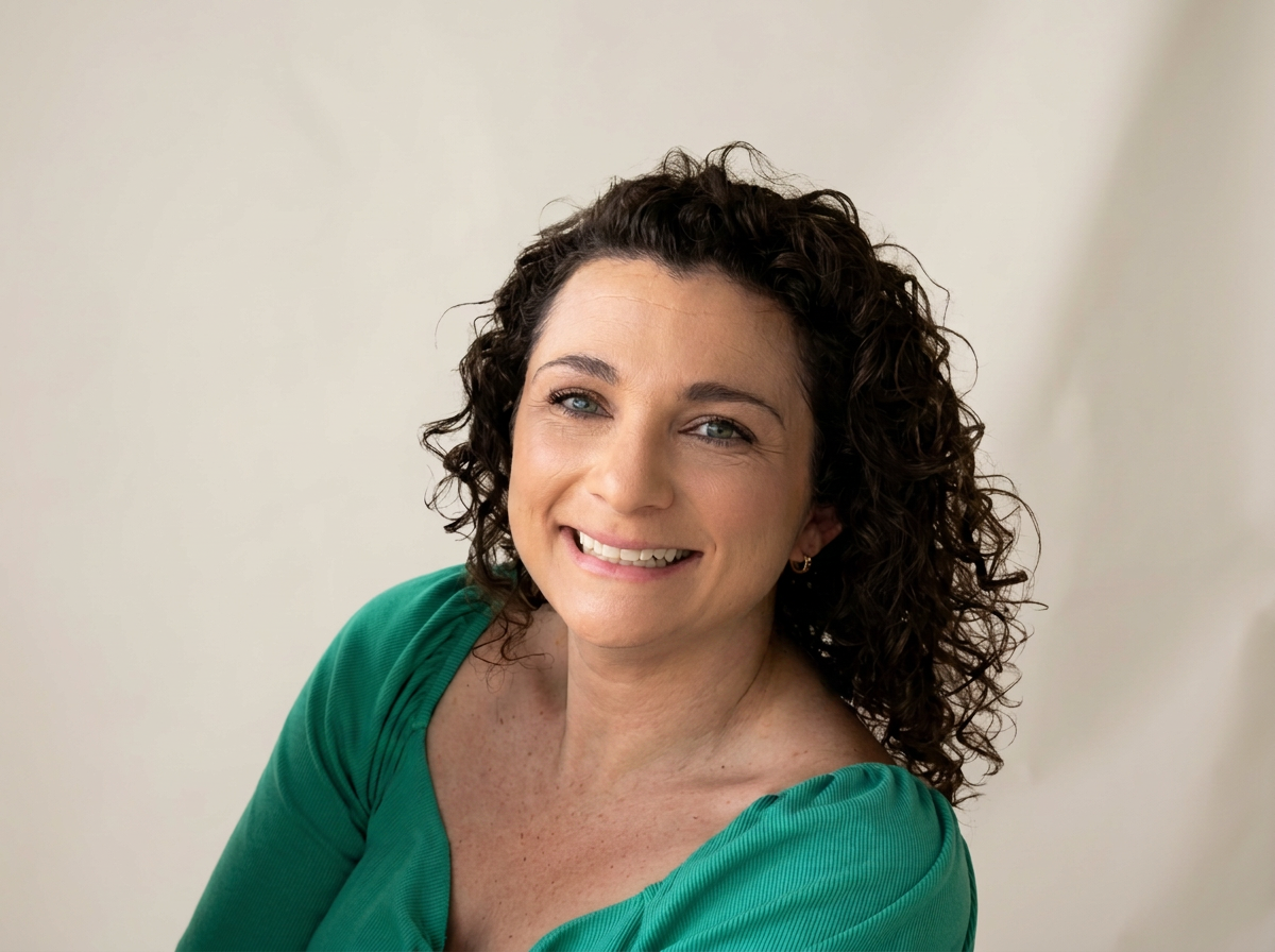 A smiling woman with curly dark hair wearing a green top against a light, neutral background.