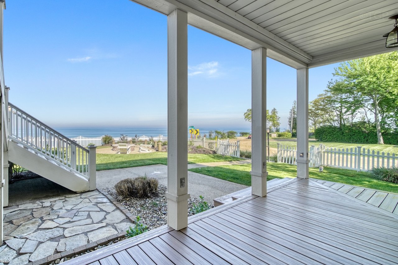 View from a porch overlooking a yard with grass, trees, and a white fence, with Lake Michigan in the background and a clear blue sky.
