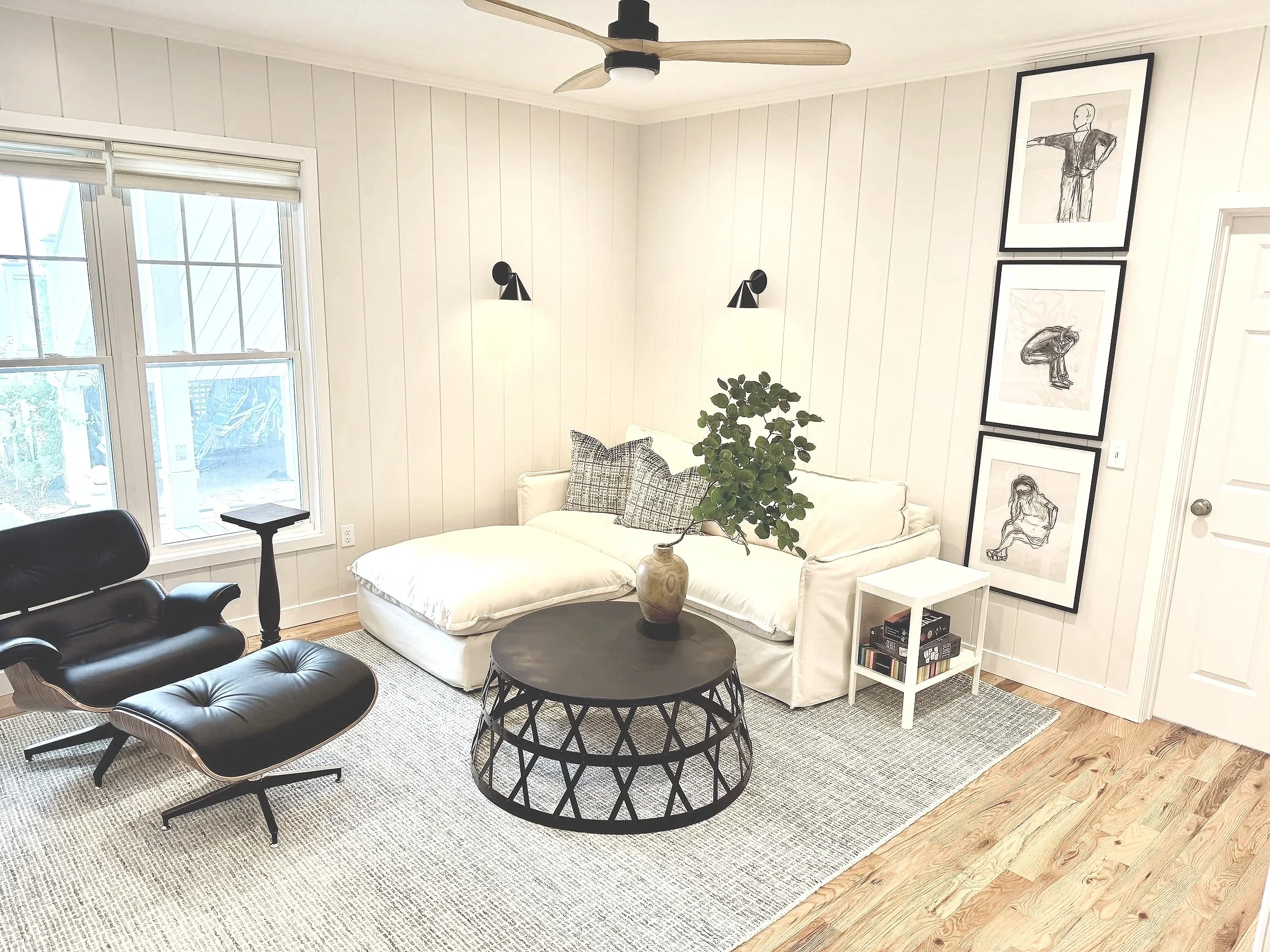 A living room with a white couch, a black leather lounge chair with a matching ottoman, a round black coffee table, a small white side table with books, a large potted plant, and wall art featuring black-and-white drawings. There are two wall-mounted lights, a ceiling fan, and a large window with blinds.