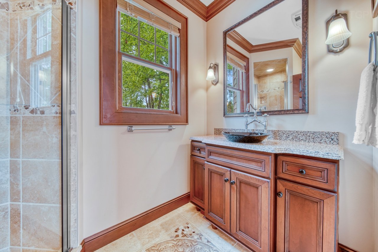 Bathroom vanity with a granite countertop, black vessel sink, and a large mirror. Wooden cabinets below, two wall-mounted light fixtures, window showing green trees outside, and part of a tiled shower on the left.