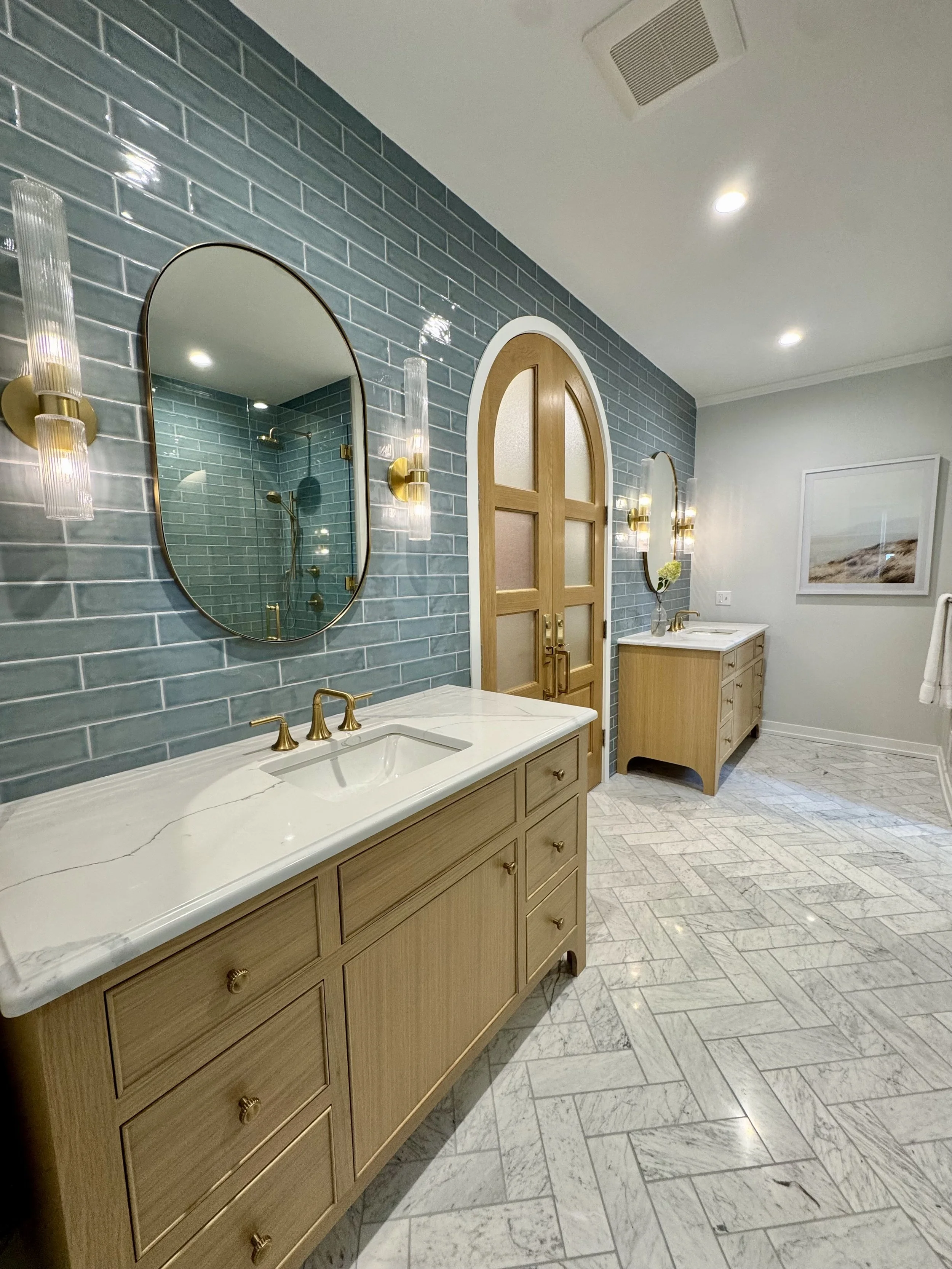 A bathroom with a light wood vanity, marble countertop, and brass fixtures. The wall behind the vanity is covered with blue subway tiles, and there are oval mirrors with brass frames and glass wall sconces. The floor has light and dark gray tiles arranged in a herringbone pattern.