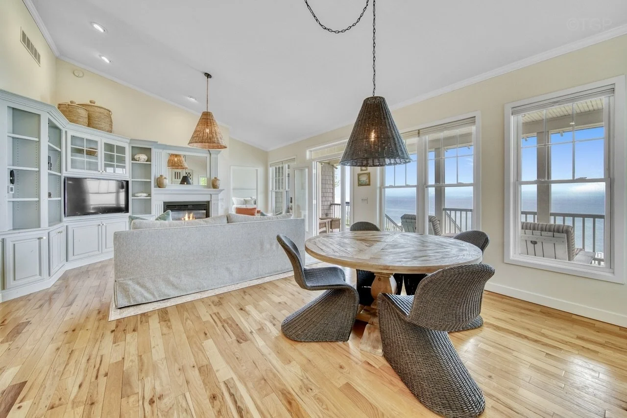 Living room with light-colored walls, hardwood floors, and large windows showing a lake view. There is a round wooden dining table with wicker chairs, a white sofa facing a built-in white entertainment center with a flat-screen TV, and a fireplace.