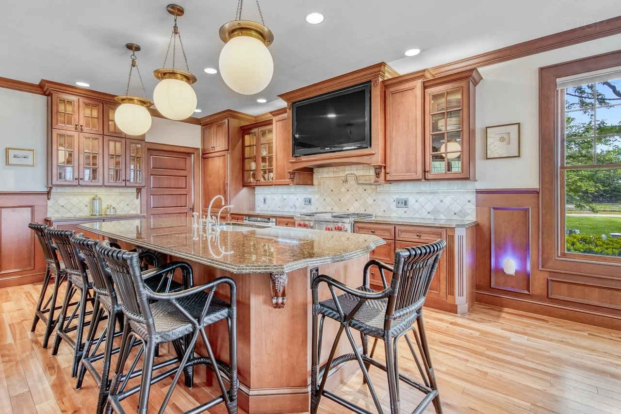 Kitchen with wooden cabinets, a granite island with a sink and bar stools, and a large window showing greenery outside.