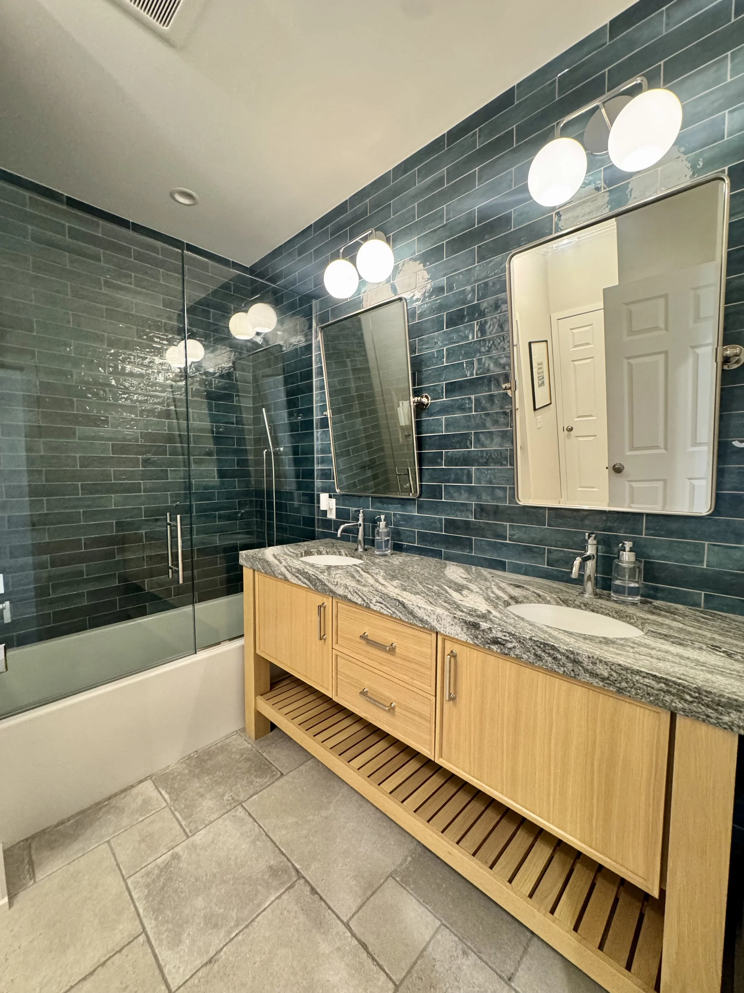 A bathroom with blue tile walls, a granite countertop vanity with two sinks and wooden cabinetry, two mirrors, modern lighting fixtures, a glass shower enclosure, and large gray floor tiles.