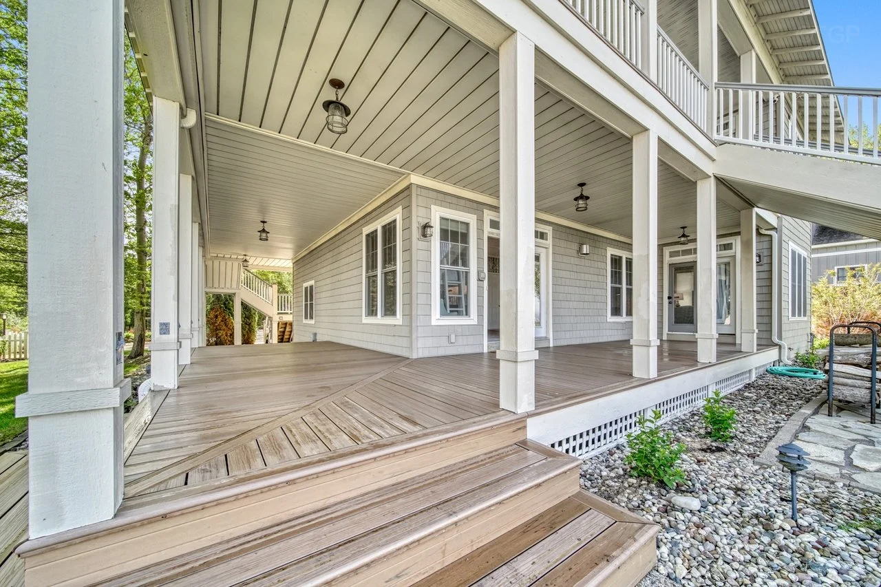 A spacious covered wooden patio with a railing, multiple ceiling lights, and steps leading down to a landscaped yard with small plants and rocks.